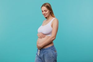 pleasant-looking-young-glad-blonde-long-haired-pregnant-woman-holding-her-belly-with-hands-smiling-positively-with-closed-eyes-isolated-blue-background